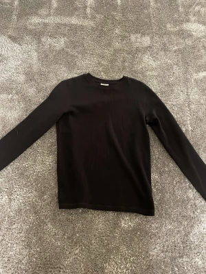 Svart ribbad långärmad tröja - Säljer en svart långärmad tröja med ribbad struktur. Tröjan har en enkel och stilren design med rund hals och är tillverkad i ett mjukt material som känns skönt mot huden. Perfekt för lager-på-lager under kyliga dagar.