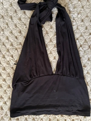 Svart omlott halterneck topp - Säljer en svart omlott halterneck topp med knyt i nacken. Toppen har djup v-ringning och är ärmlös, perfekt för att skapa en cool och trendig look. Materialet känns mjukt och stretchigt, troligen bomull eller polyester.