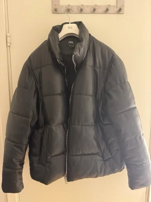 Svart pufferjacka i skinn från Zara - Säljer en svart pufferjacka från Zara med coolt quiltad design och hög krage. Jackan är tillverkad i skinnimitation och har en rejäl dragkedja framtill. Perfekt för dig som gillar en clean och modern look med lite edge.