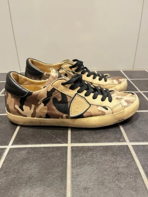 Philippe Model sneakers - Snygga sneakers från Philippe Model med beige, brunt och svart camouflage-mönster. Skorna har svarta skinnpaneler vid hälen och svarta snören. Yttermaterialet är textil och skinn, med en platt sula i gummi. Perfekt för dig som gillar streetstyle och unika detaljer. Storlek IT 41 = EU 42. 