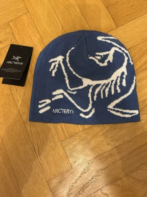 Blå mössa från Arc'teryx - Snygg blå mössa från Arc'teryx med vit grafisk logga på framsidan. Mössan är stickad och har en enkel, rundad form som sitter skönt på huvudet. Perfekt för kalla dagar och outdoor-aktiviteter. Materialet är mjukt och värmande.
