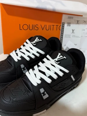 Louis Vuitton svarta sneakers (HELT NY) - Säljer ett par svarta Louis Vuitton sneakers med vita snören och tydliga LV-loggor på både plös och sidor. Skorna har präglade monogramdetaljer, robust sula och coola kontraster i svart och vitt. Tillverkade i skinn och syntetmaterial för en lyxig känsla.