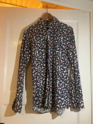 Blommig skjorta från Eton, strl 39 - Snygg skjorta från Eton med blommigt mönster i färger som lila, röd, vit och orange på en mörkblå botten. Skjortan har lång ärm, klassisk krage och knappar framtill. Perfekt för dig som vill sticka ut med en cool och färgglad stil.