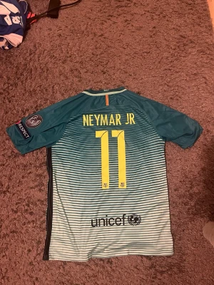 Neymar 16/17 tredjeställ fotbollströja - Barcelona fotbollströja med Neymar Jr och nummer 11. Tröjan är randig i turkos och vit med gula detaljer och Champions League-märke på ärmen.