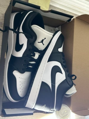 Nike Air Jordan 1 Low svart/vit - Nike Air Jordan 1 Low sneakers i svart och vitt med klassisk Swoosh på sidan och Jumpman-logga på tungan. Skorna har snörning, rund tå och platt sula. Tillverkade i skinn med perforeringar på tån för extra ventilation. Perfekt för dig som gillar stilrena och ikoniska sneakers.