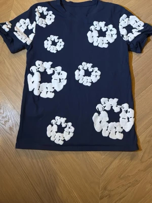 Mörkblå t-shirt med vita blommor - Säljer en mörkblå t-shirt med coola vita blommönster över hela plagget. T-shirten har korta ärmar och är gjord i mjuk bomull som känns skön mot huden. Perfekt för dig som gillar unika prints och vill sticka ut lite extra.