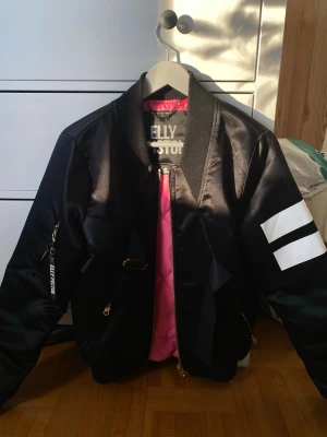 Svart bomberjacka från Elly Pistol - Snygg svart bomberjacka från Elly Pistol med coola vita detaljer på ärmen och rosa foder. Jackan har guldfärgade dragkedjor, ribbad krage och flera fickor. Perfekt för dig som vill sticka ut med en unik streetstyle-look.