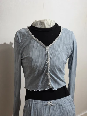  pyjamas set med cardigan och byxor - Ett sött set bestående av en cardigan med knappar och långärmar, samt ett par matchande byxor. Cardigan har en vågig kant med spetsdetaljer längs knapparna och halsringningen. Byxorna har en resår i midjan och en dekorativ rosett