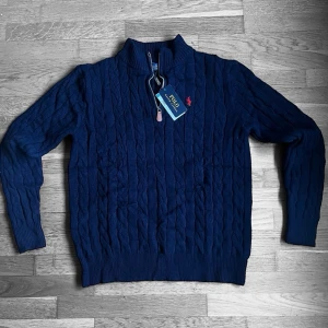 Marinblå stickad half zip från Polo Ralph Lauren - Säljer en marinblå stickad tröja från Polo Ralph Lauren med half zip och kabelstickat mönster. Tröjan har lång ärm, ribbade muddar och röd broderad logga på bröstet. Perfekt för dig som gillar klassisk och stilren design.