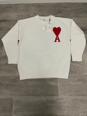 Vit stickad tröja från Ami - Vit stickad tröja från Ami med rött hjärta och bokstaven A broderat på bröstet. Tröjan har rund hals, ribbade muddar och nederkant samt långärmad passform. Perfekt för dig som gillar stilrena detaljer och vill ha något som sticker ut lite.