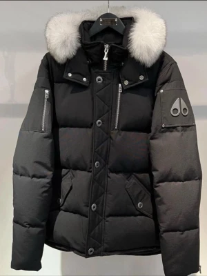 Svart Moose Knuckles pufferjacka med päls - Svart pufferjacka från Moose Knuckles med vit fluffig päls på huvan, silverfärgade dragkedjor och knappar. Jackan har flera fickor, ikonisk logga på ärmen och en snygg, modern look. Perfekt för kalla dagar och riktigt street.