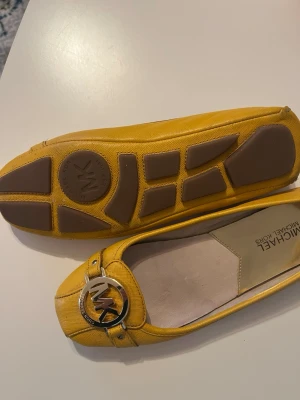 Gula ballerinaskor Michael Kors - Snygga gula ballerinaskor från Michael Kors med rund tå och platt sula. Skorna har en metallfärgad MK-logga framtill och insida i guld. Yttermaterialet är skinn med en lätt textur. Perfekt för dig som vill ha en färgklick till outfiten.