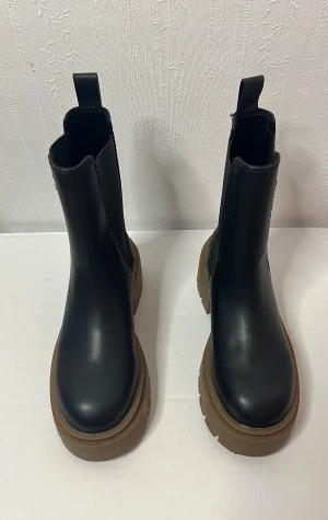 Svarta chelsea boots från H&M - Säljer ett par svarta chelsea boots från H&M med chunky brun sula och rund tå. Bootsens ovandel är i syntetmaterial och har elastiska paneler på sidorna för enkel påtagning. Perfekt för dig som gillar robust stil och vill ha något som sticker ut. Har otur med att ha små men breda fötter..