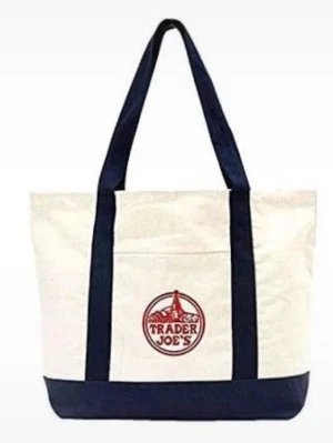 Trader Joe's shoppingväska i bomull - Säljer en stor och praktisk shoppingväska från Trader Joe's i vit och mörkblå bomull. Väskan har långa handtag och en framficka med röd logga. Perfekt för att bära med sig allt man behöver till skolan, stranden eller stan.