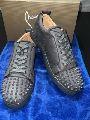 Christian Louboutin svarta sneakers med nitar - Svarta sneakers från Christian Louboutin med mattsvart mocka och coola nitar på tån. Skorna har snörning och den klassiska röda sulan med logotyp. Insidan är beige och de har en rund tå. Perfekt för dig som vill sticka ut med en edgy stil.