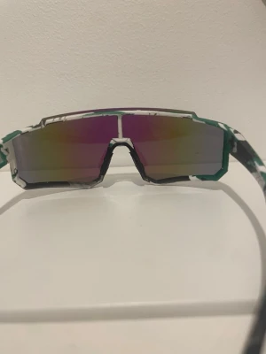 Sol glasögon från Cypern  - Bilderna visar ett par sportiga solglasögon med spegelglas i blå och gröna toner samt vit och svart mönstrad båge. Ingen väst syns på bilderna.