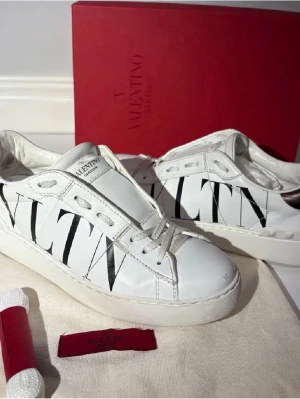 Valentino Garavani Open sneakers - Snygga vita Valentino Garavani Open sneakers med svart VLTN-tryck på sidorna och metallic-silver detalj på hälen. Skorna har snörning, rund tå och platt sula. Tillverkade i skinn med coola nitar bak på sulan. Perfekt för dig som gillar lyxiga street vibes.