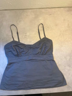  linne vintage Hollister XS - Säljer en mörkgrå linnetopp från Hollister i storlek XS. Toppen har justerbara smala axelband, v-ringad hals med dekorativa knappar framtill och lätt rynkade detaljer vid bysten. Materialet är mjukt och känns som bomull, perfekt för varma dagar.