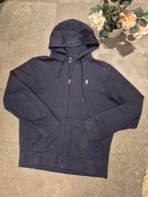 Marinblå ralph lauren hoodie - * 👕 Marinblå hoodie från Ralph Lauren * ✨ Mycket bra skick, sparsamt använd * 🕶️ Klassisk och tidlös design * 🔒 Dragkedja framtill och huva med snörning * 👌 Passar till många olika tillfällen – både vardag och mer uppklätt * 👖 Lätt att matcha med jeans, chinos eller mjukisbyxor * 📏 Storlek XS, men passar som S * ⭐ Hög kvalitet och bekväm passform 