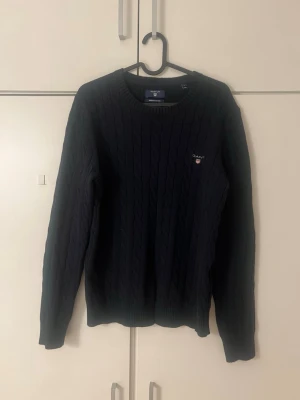 Mörkblå stickad tröja från GANT - Säljer en mörkblå stickad tröja från GANT med klassiskt kabelstickat mönster och rund hals. Tröjan har lång ärm och liten broderad logga på bröstet.