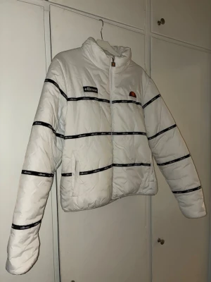 Vit pufferjacka från Ellesse - Säljer en vit original pufferjacka från Ellesse med svarta ränder och logotape längs ärmar och kropp. Jackan har hög krage, dragkedja framtill och broderad Ellesse-logga på bröstet. Materialet är syntet och jackan är quiltad för extra värme. Perfekt för kalla dagar.