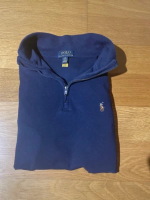 Marinblå half zip tröja Polo Ralph Lauren - Snygg marinblå tröja från Polo Ralph Lauren med klassisk broderad logga på bröstet. Modellen har half zip-dragkedja och hög krage. Tillverkad i mjuk bomull som känns skön mot huden. Perfekt för dig som gillar stilrena och tidlösa plagg.
