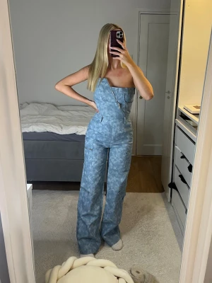 Blå byxdress från Mars the Label - Säljer en blå byxdress från Mars the Label i storlek 8. Byxdressen har ett coolt mönster, breda ben och en sned dragkedja framtill. Modellen är strapless och har fickor på benen för extra stilpoäng. Perfekt för dig som vill sticka ut med en unik look.