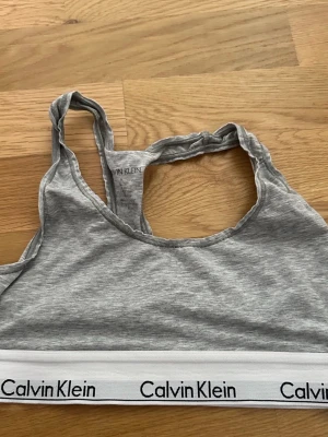 Bralette  från Calvin Klein - Grå Calvin Klein sport-bh med vit resårkant med märkets namn tryckt på. Bh:n har en racerback-design.  Det enda anmärkningsbara är att tvättrådslappen är avklippt då den skavde, i övrigt inget att anmärka på :)
