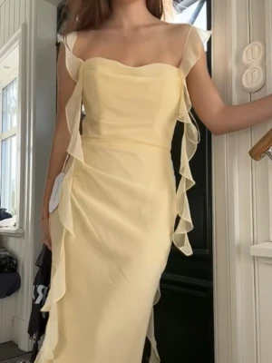 Ljusgul långklänning med volanger - Säljer en elegant ljusgul långklänning med transparenta axelband och dekorativa volanger längs sidorna. Klänningen har en figurnära passform och är tillverkad i ett tunt, flowy material som ger en romantisk känsla. Perfekt för speciella tillfällen.