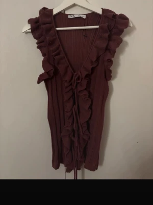 Vinröd ribbad topp med volanger från Zara - Säljer en vinröd ribbad topp från Zara med snygga volanger längs v-ringningen och framtill. Toppen är ärmlös och har knytband framtill för extra detalj. Perfekt att styla med jeans eller kjol för en trendig look.
