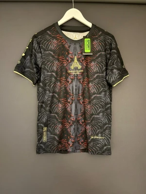 Svart och röd fotbollströja S - Säljer en svart och röd fotbollströja från Comma Football med coolt abstrakt mönster och guldfärgade detaljer. Tröjan har korta ärmar, rund hals och texten 'European Champions 31.05.2025' med Eiffeltornet broderat på bröstet. Perfekt för dig som gillar unik design.