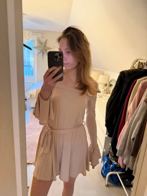 Ljusrosa offshoulder kortklänning - Supersöt ljusrosa kortklänning med offshoulder och långa trumpetärmar. Klänningen har ett knytband i midjan som ger en snygg siluett och är gjord i ett mjukt, stretchigt material. Perfekt för dig som gillar en feminin och trendig look.