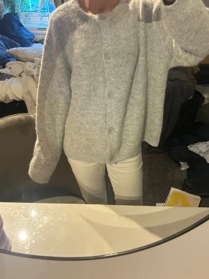 Ljusgrå stickad kofta med knappar - Mysig ljusgrå stickad kofta med rund hals och stora knappar framtill. Koftan har en loose passform och långa ärmar, perfekt för att hålla sig varm under kyliga dagar. Materialet är fluffigt och mjukt, vilket gör den extra skön att bära.