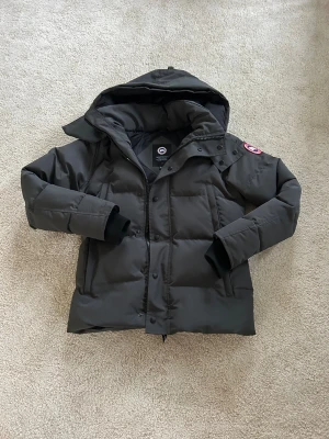 Canada Goose Jacka - Snygg mörkgrå dunjacka från Canada Goose med huva och ikonisk röd-vit logga på ärmen. Jackan har långa ärmar, dragkedja och knappar framtill samt två sidofickor. Perfekt för kalla dagar och har en clean, stilren look som funkar till streetwear.