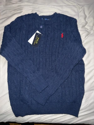 Blå stickad tröja från Polo Ralph Lauren - Säljer en marinblå stickad tröja från Polo Ralph Lauren i storlek S. Tröjan har kabelstickat mönster, rund hals och långärmade ribbade muddar. På bröstet finns den klassiska röda Polo-loggan broderad. Perfekt för dig som gillar stilrena och tidlösa plagg.