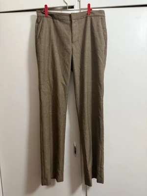 Rutig kostymbyxa från Ralph Lauren - Snygga rutiga kostymbyxor från Lauren Ralph Lauren i beige och bruna toner. Klassisk rak passform med midwaist och diskret mönster. Perfekt för dig som gillar stilrena och tidlösa plagg. Materialet är mjukt och bekvämt, med fickor på sidorna.