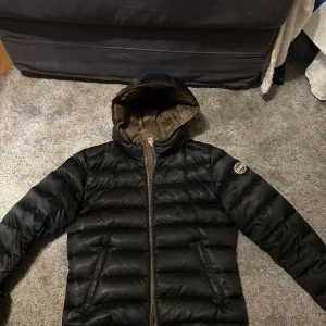 Svart pufferjacka från Colmar - Snygg svart pufferjacka från Colmar med brunt foder och huva. Jackan har quiltad design, dragkedja framtill och två fickor. Colmar-logga på ärmen ger en clean och sportig vibe. Perfekt för kalla dagar när du vill hålla stilen.