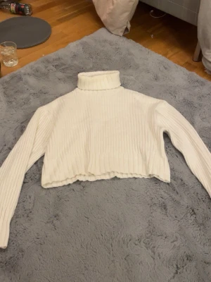 Vit croppad polotröja i stickad ribb - Säljer en vit croppad polotröja med ribbad stickning. Tröjan har lång ärm och hög polokrage, perfekt för att hålla värmen. Den är gjord i ett mjukt stickat material och har en modern, loose passform som är både bekväm och snygg. Kan eventuellt tänka mig gå ner i pris