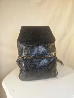 Svart ryggsäck i skinn med gulddetaljer - Snygg svart ryggsäck i skinn med justerbara axelremmar och flera praktiska fack. Väskan har guldfärgade dragkedjor och spännen som ger en cool touch. Perfekt storlek för att få plats med det du behöver och passar till många olika stilar.