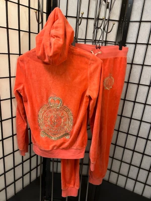 Juicy Couture velour tracksuit – komplett set, Y2K, coral/orange - Säljer ett original Juicy Couture velour-set bestående av hoodie och matchande byxor i korall/orange. Typisk Y2K-stil med klassisk Juicy-logga prydd med dekorativa detaljer. Glam tracksuit Perfekt för dig som gillar 2000-talsmode, comfy outfits eller vill ha en statement-tracksuit.  Storlek: Large Skick: Bra skick för sin ålder! Saknas en detalj på insidan av ena knappen på byxfickan och lappen på byxans insida sitter löst.  Material: Mjuk velour  ✔ Komplett set ✔ Ikonisk Juicy Couture-stil ✔ So