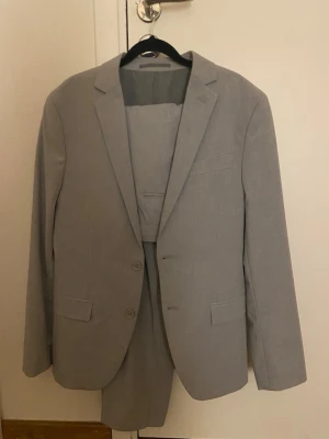 Ljusgrå kavaj och kostymbyxa - Snygg ljusgrå kostym med klassisk kavaj och matchande kostymbyxa. Kavajen har två knappar framtill, fickor med lock och en stilren krage. Materialet känns lätt och är perfekt för dig som vill ha en clean och modern look.