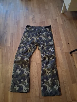 Camouflage täckbyxor från Slade Couture - Säljer ett par feta täckbyxor från Slade Couture i storlek S. Byxorna har camouflage-mönster i gröna och bruna toner, flera fickor med lock och slitstark polyester. Perfekta för kalla dagar och outdoor-aktiviteter. Snygg logga på benet.