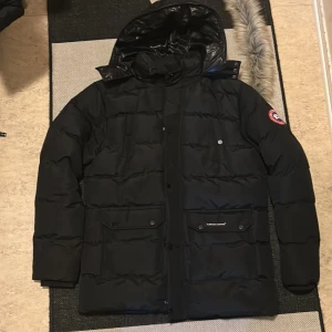 Canada Goose Jacka - Säljer en svart dunjacka från Canada Goose i storlek L. Jackan har huva, flera fickor med lock och tryckknappar samt klassisk Canada Goose-logga. Päls medföljer
