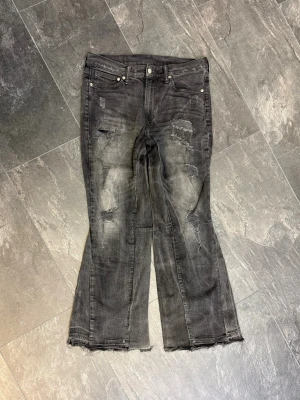 MARES TAKEOVER 1 OF 1 ”MIDNIGHT” - Bootcut jeans i svart tvättad denim , med slitna detaljer och råa kanter vid bensluten. Jeansen har fem fickor, knappgylf och coola slitningar framtill för en edgy vibe. Passar dig som gillar street och unik stil.