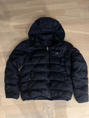 Svart pufferjacka från GANT mörk blå - Svart pufferjacka från GANT med huva och dragkedja framtill. Jackan har quiltad design, två sidofickor med dragkedja och en diskret logga på bröstet. Perfekt för kyliga dagar och har en snygg, enkel look som funkar till det mesta. Ny pris 2200 jackan är mörk blå 
