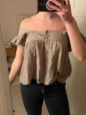 Beige offshoulder blus med knappar - Säljer en beige blus med offshoulder och korta volangärmar. Blusen har en luftig passform och knappar framtill. Materialet är lätt och skrynkligt, vilket ger en avslappnad vibe. Perfekt för varma dagar och enkel att styla med jeans eller kjol.
