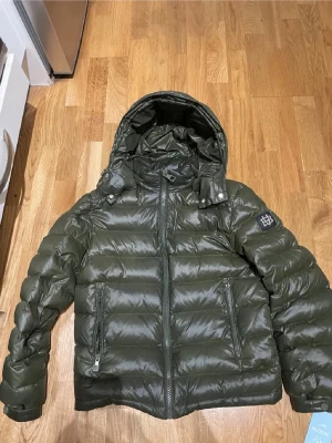 Grön pufferjacka från Bvaldi XS - Säljer en grön pufferjacka från Bvaldi i storlek XS. Jackan har huva, två fickor med dragkedja och ett snyggt Bvaldi-märke på ärmen. Den är quiltad och har en glansig yta som ger en cool look. Perfekt för kalla dagar och enkel att matcha med jeans.