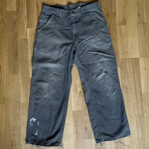 Grå/Brun Carhartt cargopants  - Säljer ett par grå carhartt byxor med bred passform och flera fickor, inklusive bakfickor med patchdetalj. Byxorna har råa, fransiga benslut och några vita färgstänk på benen för en cool, sliten look. Dem är riktigt bra passform. Jag är 1.78cm och de sitter riktigt bra på mig. Färg har kommit på dem. Själv tycker jag de gör dem bättre men ni får avgöra själva. Bredd: 40cm Längd: 91cm. Skriv för frågor. Pris kan diskuteras.
