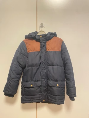 Mörkblå pufferjacka Hampton Republic - Snygg mörkblå pufferjacka från Hampton Republic med bruna axelpartier och huva. Jackan har guldiga knappar, två stora fickor framtill och broderad logga på ärmen. Perfekt för kalla dagar och har en schysst, lite sportig vibe.