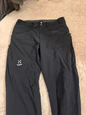 Svarta täckbyxor från Haglöfs - Svarta täckbyxor från Haglöfs med två knappar i midjan och dragkedjor vid fickorna. Byxorna har en rak passform och är gjorda i slitstarkt syntetmaterial, perfekta för utomhusaktiviteter. Loggan syns tydligt på ena benet.